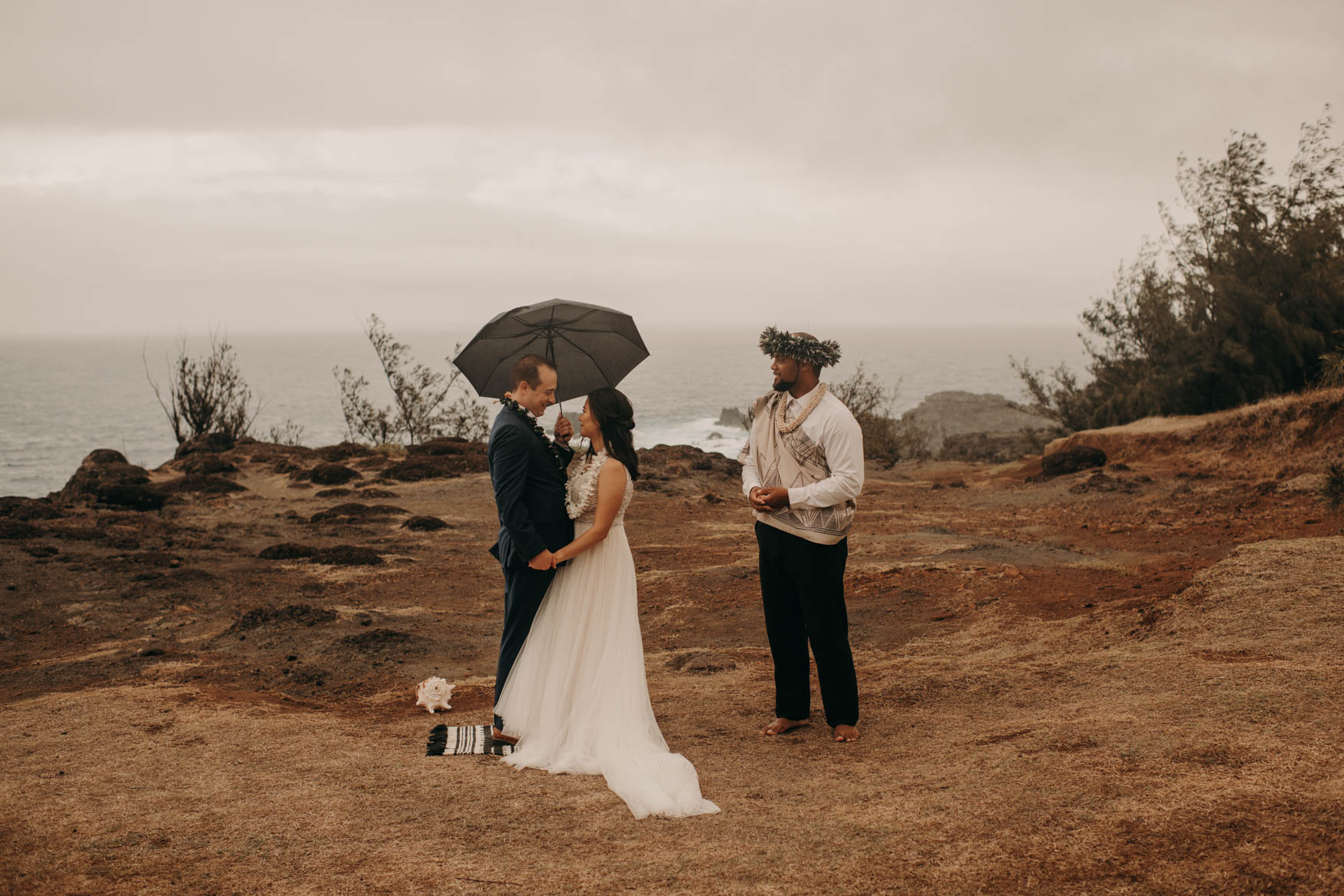 Andrew and Alison stand closely together under an umbrella andrew is holding in his left hand. Euta looks at them with his hands held together and speaking.