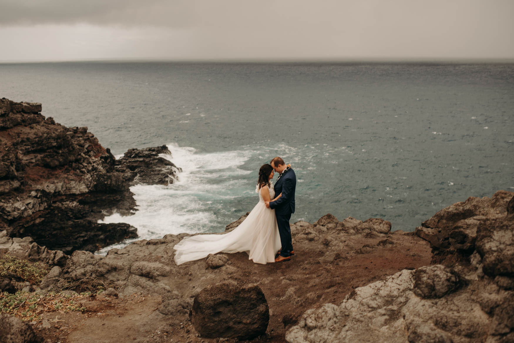 Andrew and Alison stand together on a cliff, the grey-blue ocean crashing on rocks behind and below them.