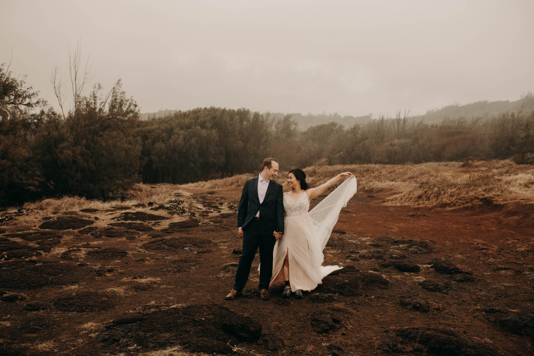 Andrew and Alison stand together in a red clay field with patches of yellow dry grass, mist rolling in, and green shrubs in the further background. They hold hands. Andrew looks at Alison, smiling. Alison looks at Andrew, smiling, while holding the train of her dress up with her left hand.