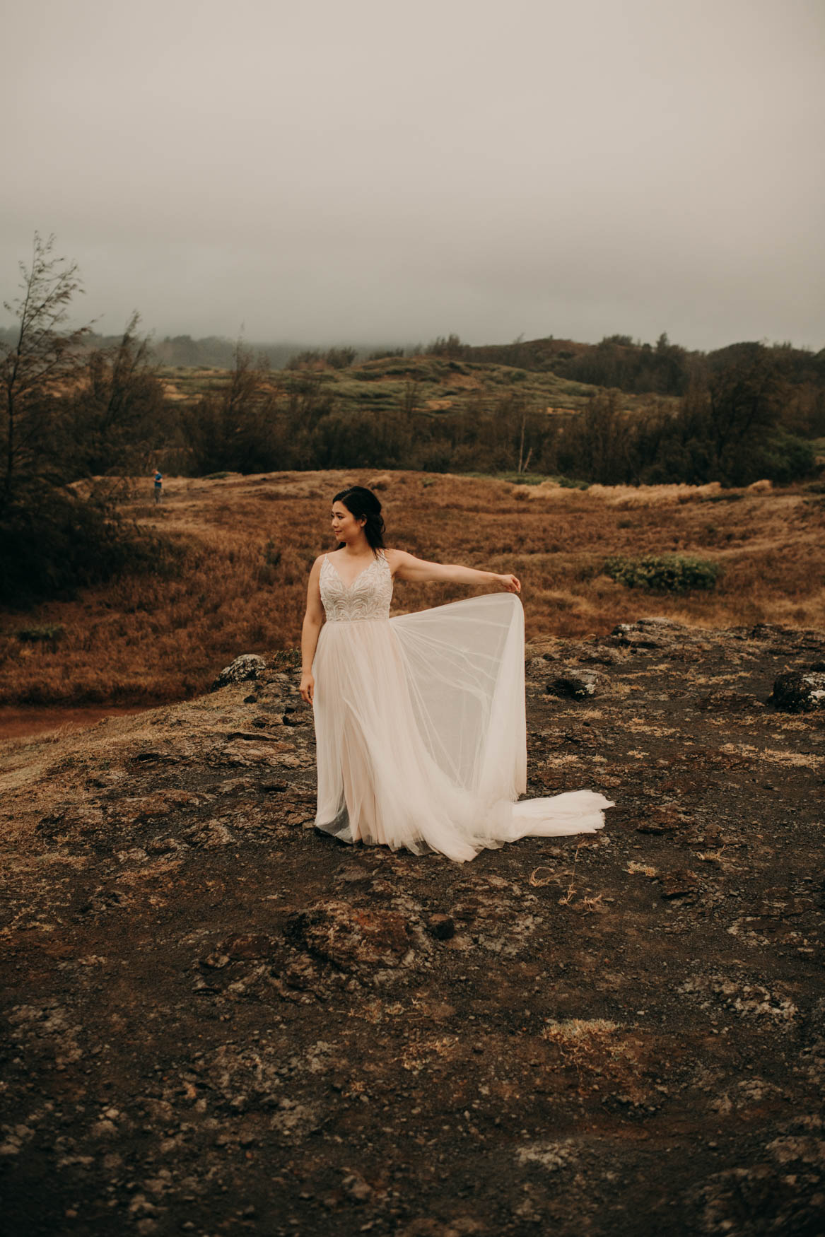 Alison stands alone on a rocky bluff, a feild of yellow grass in the background, and green trees and hills in the further background. She holds her dress up with her left hand, allowing the light to shine through the train. She looks off into the distance to her right.